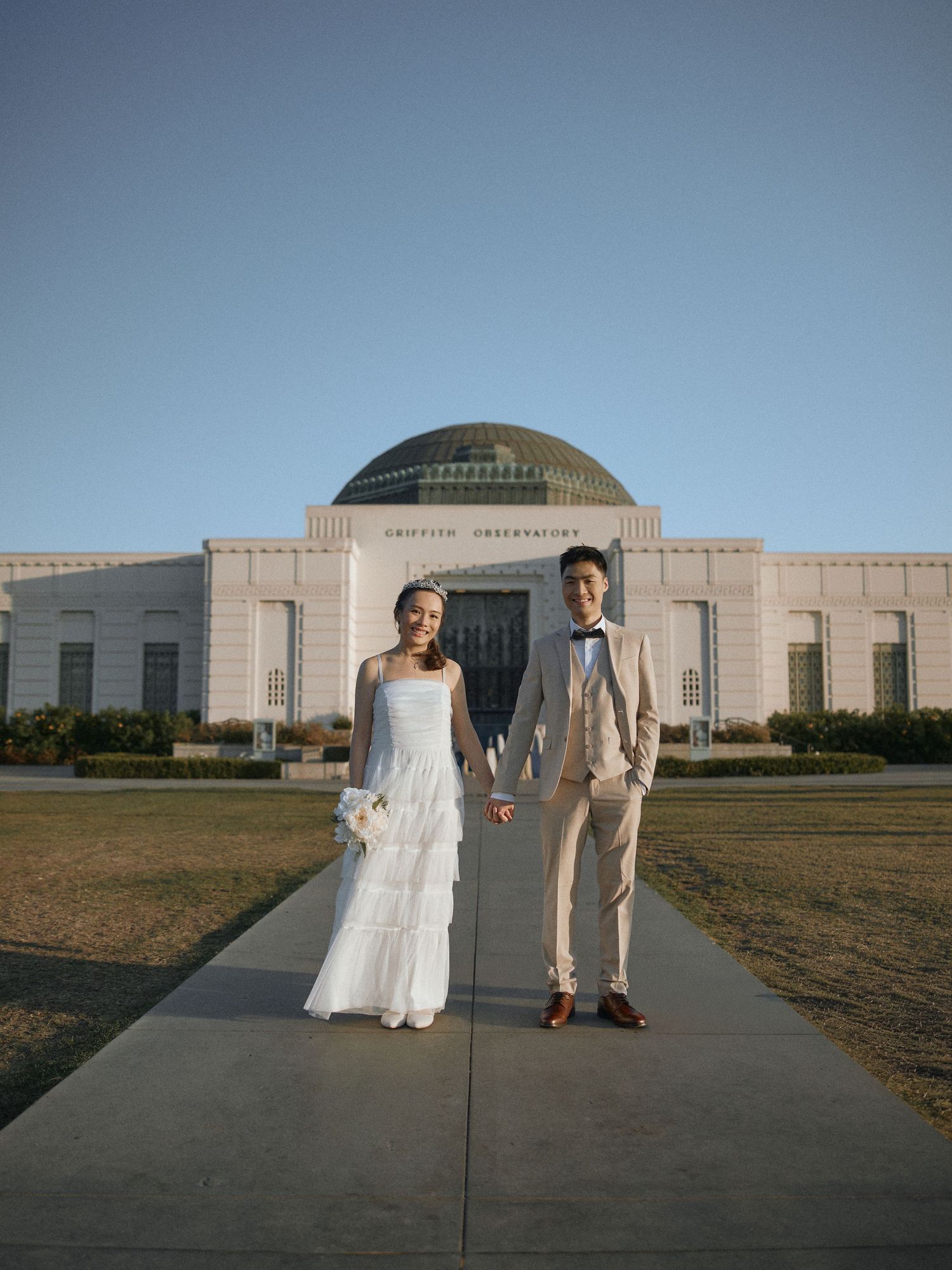 Couple holding hands walking in front of the iconic white Griffith Observatory dome at sunrise in Los Angeles.