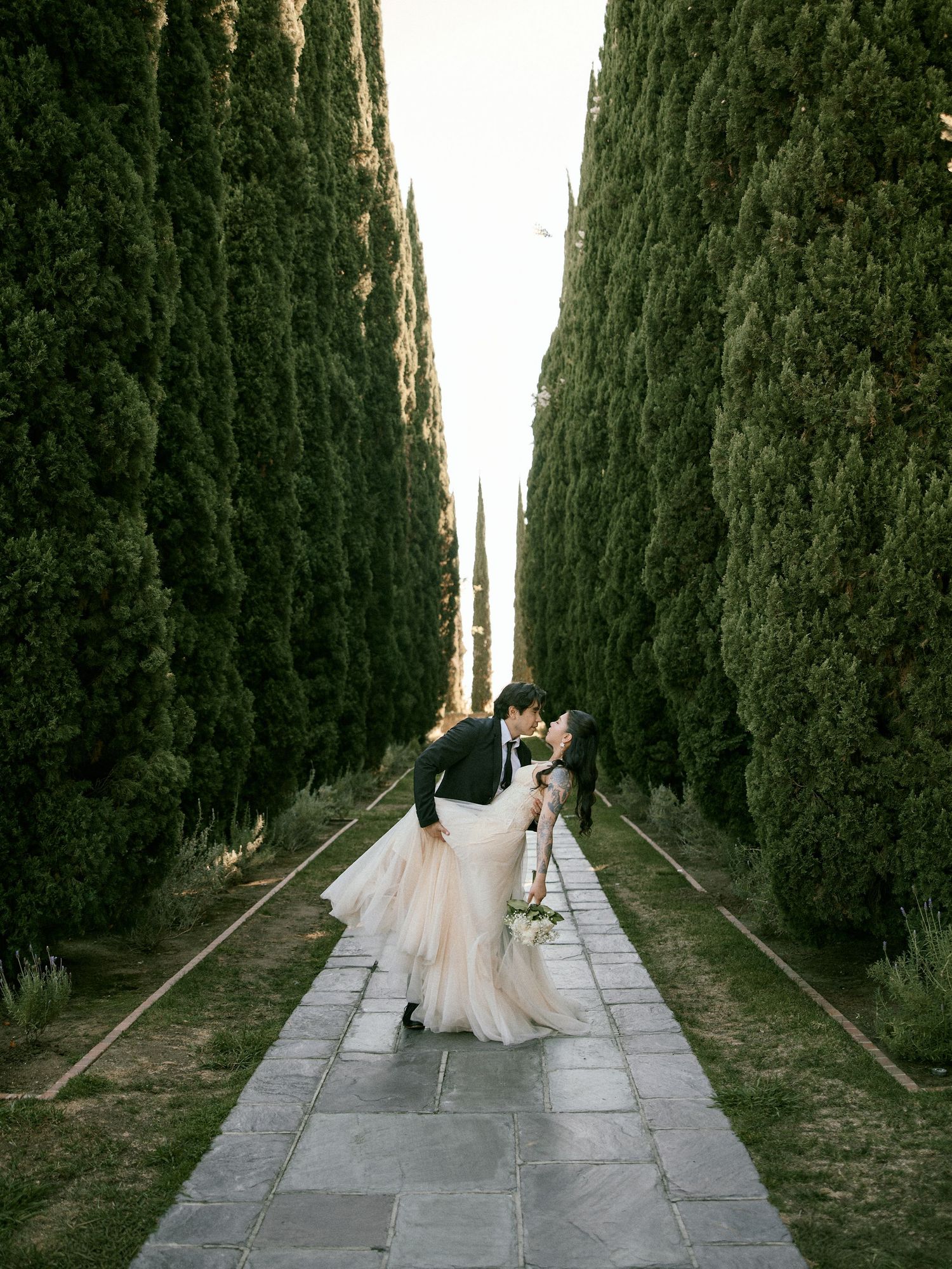 Engaged couple walking hand-in-hand down a scenic stone pathway lined with tall cypress trees, captured in a romantic, candid style.