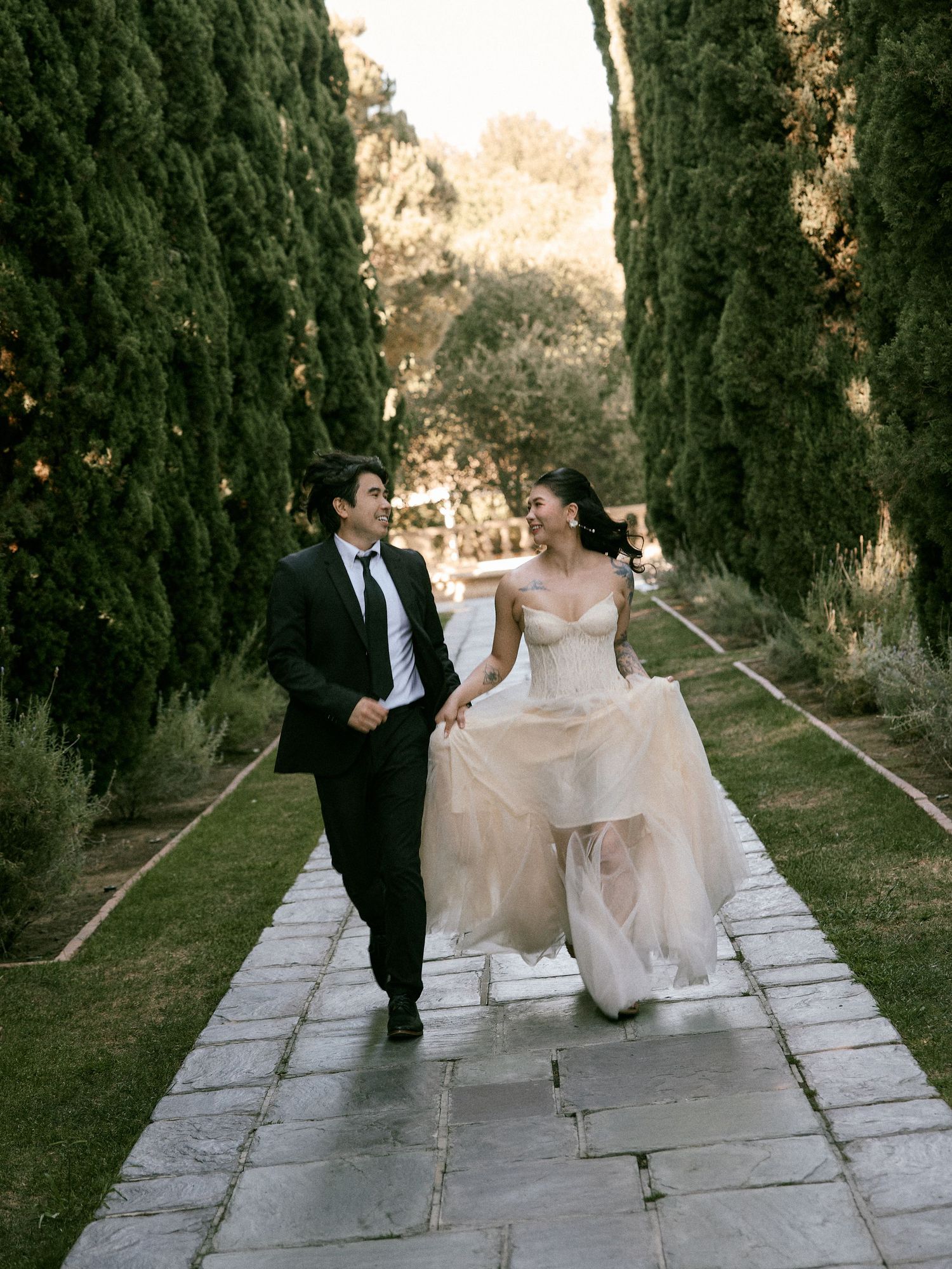 Bride in an elegant gown and groom walking together along a stone walkway in a scenic outdoor setting, blending digital and film aesthetic