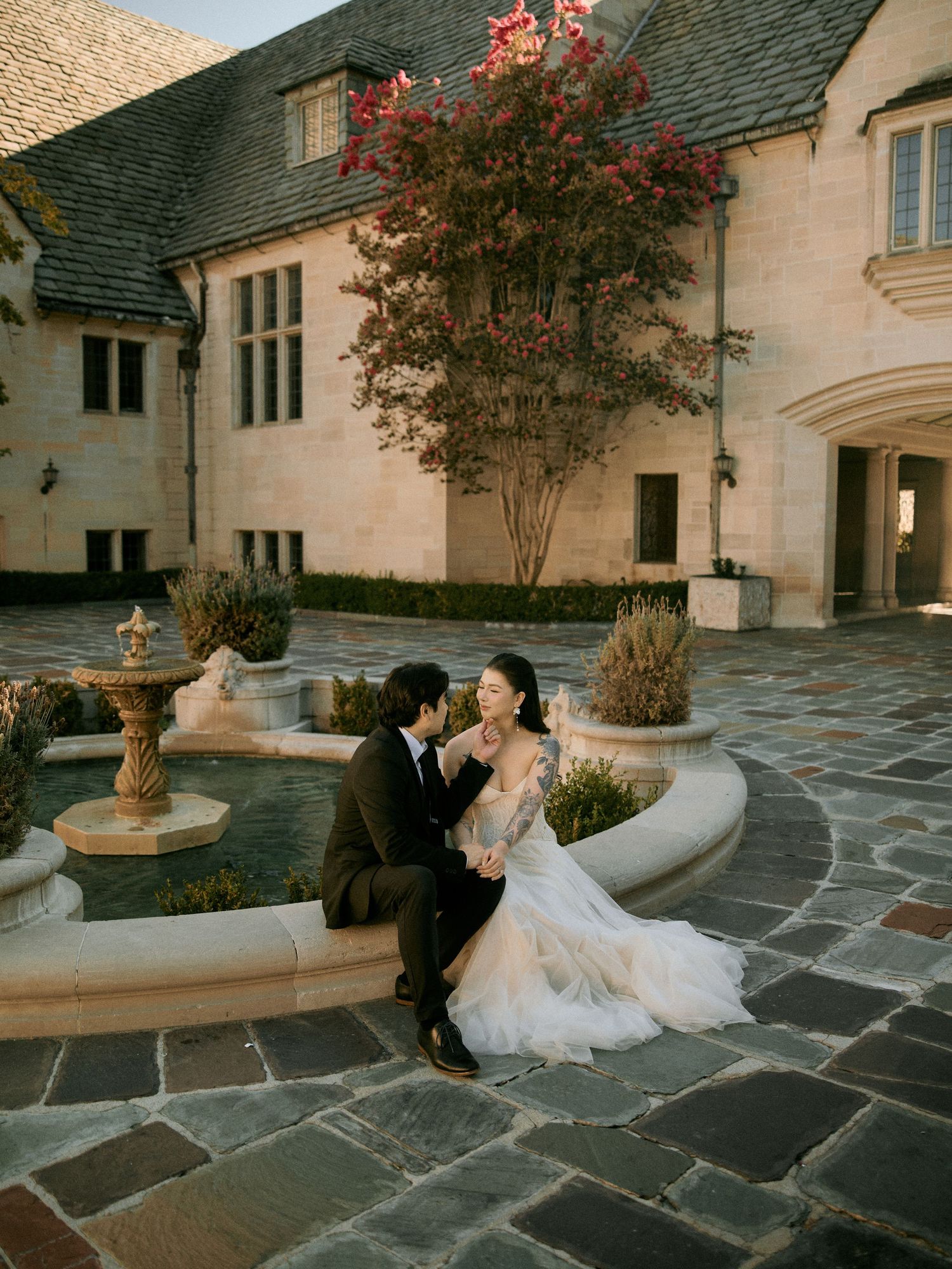 Bride and groom sitting closely on a stone fountain ledge outside a classic architectural estate during a wedding and engagement photo shoot
