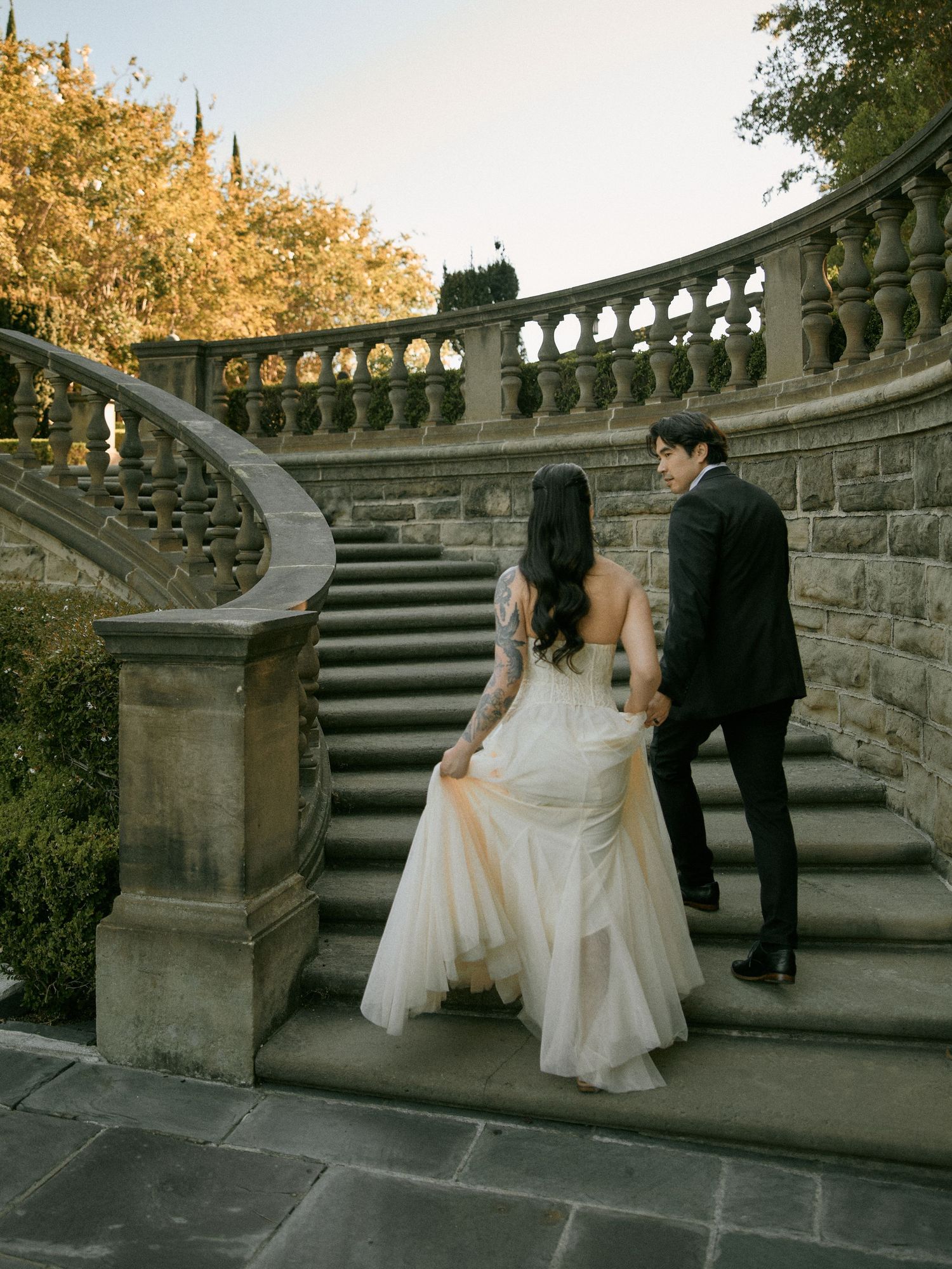 Bride and groom walking up ornate outdoor stone steps surrounded by lush green foliage during an engagement photo session.
