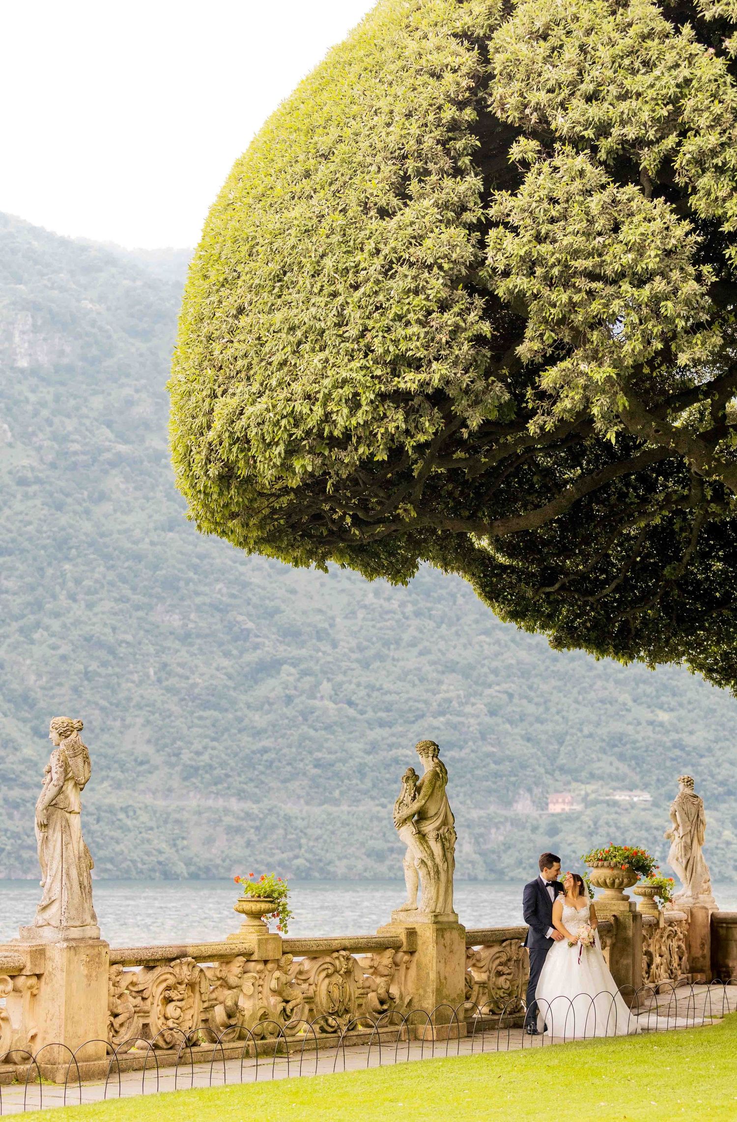 Romantic couple embraces under a manicured tree at Villa del Balbianello overlooking Lake Como in Italy.