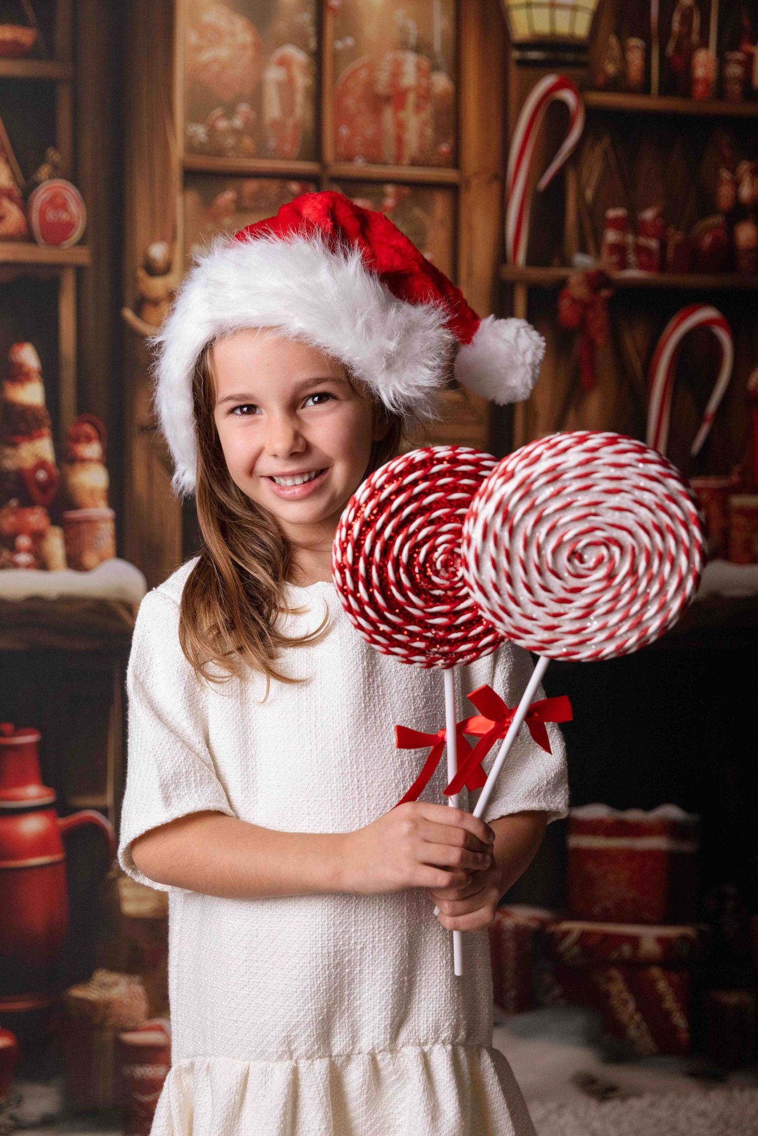 Young child in white dress holding large red and white striped lollipop decorations in festive Christmas setting.