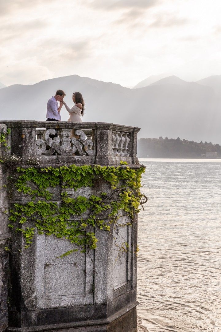 Romantic couple on ivy-covered stone balcony overlooking misty lake with mountain backdrop at dusk.