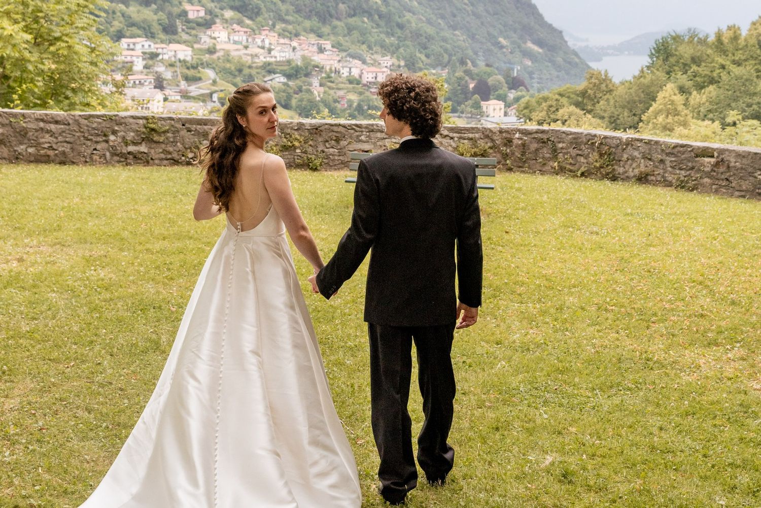 A couple in wedding attire holds hands while walking on a grassy lawn overlooking a scenic mountain landscape.