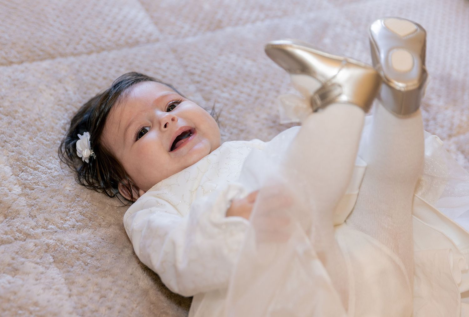 Baby in white outfit lying on back on quilted surface with happy expression.