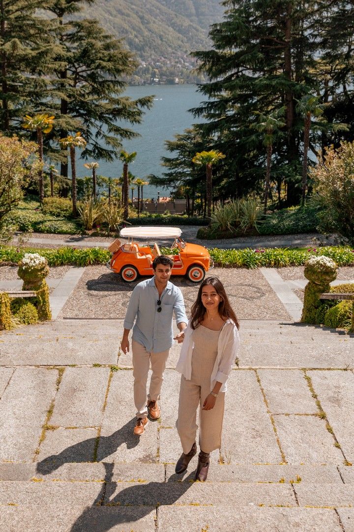 Couple walking hand-in-hand in an Italian garden with a vintage orange car and Lake Como in the background.