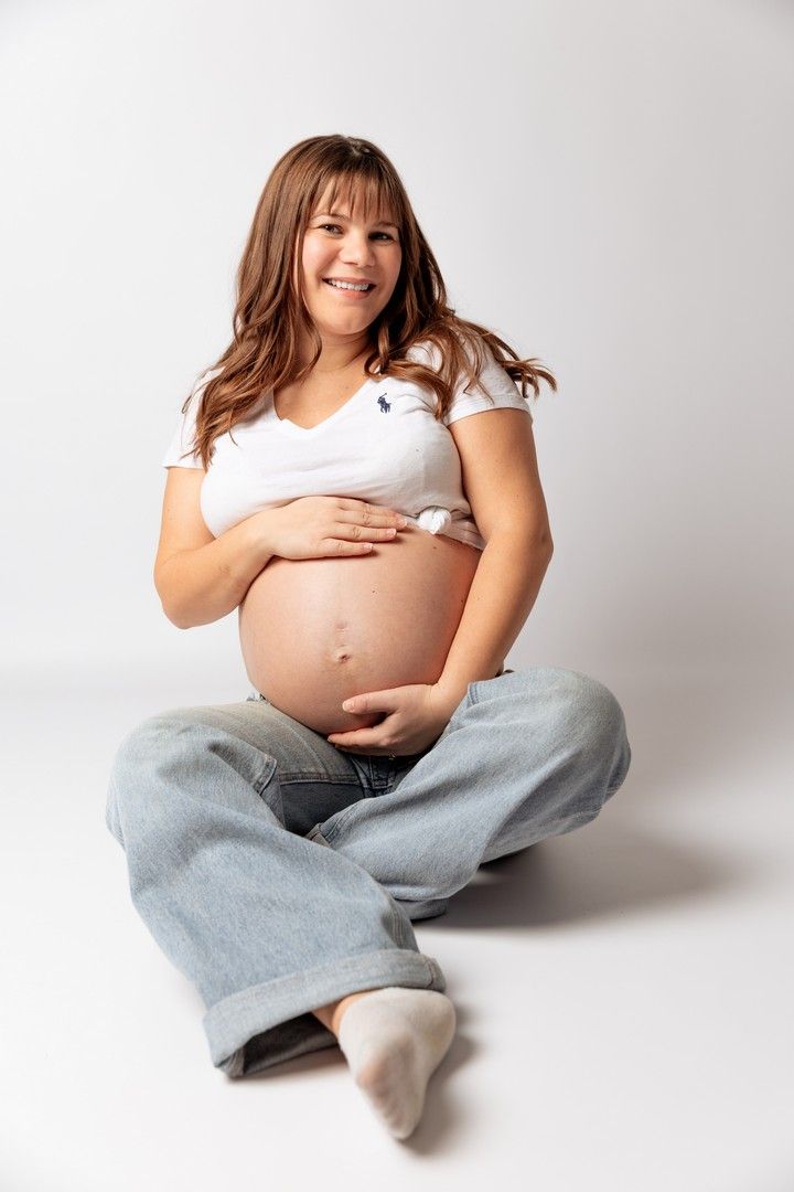 Smiling pregnant woman sitting cross-legged on floor holding her bare belly in studio.