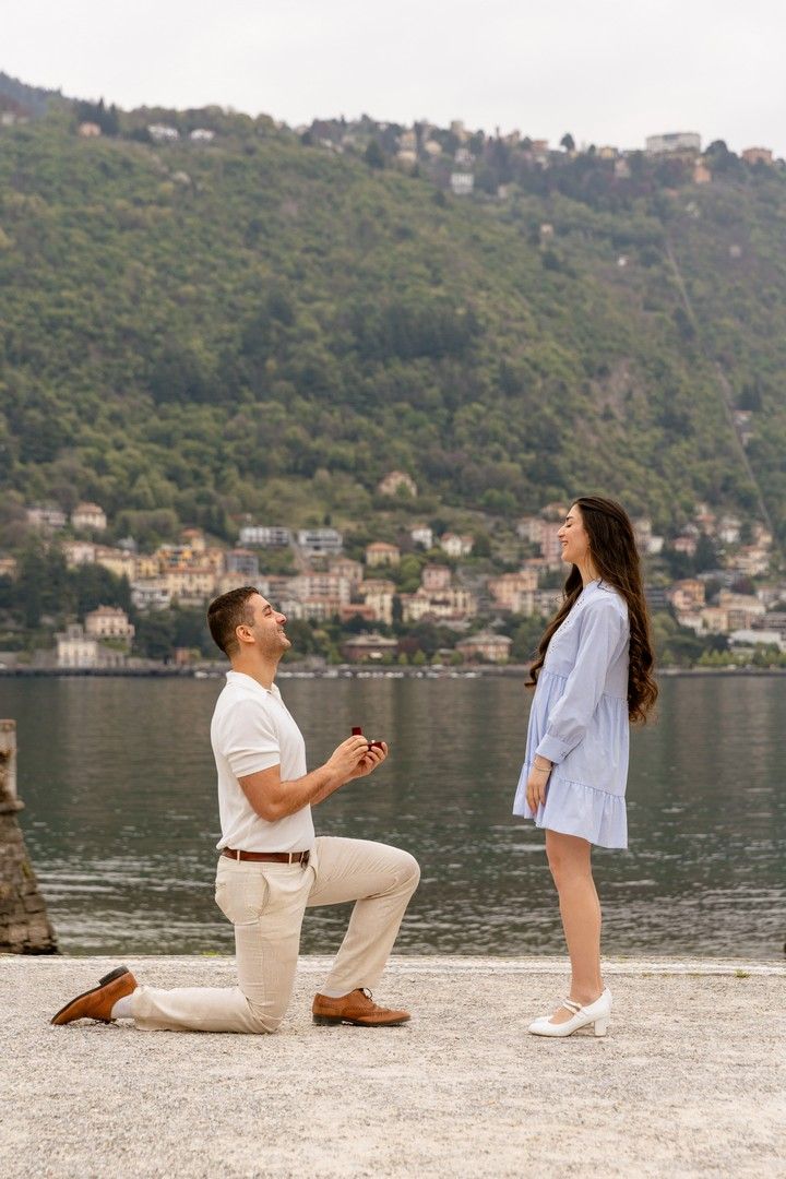 Man proposing on one knee to woman by Lake Como, Italy, with scenic mountains and waterfront village backdrop.