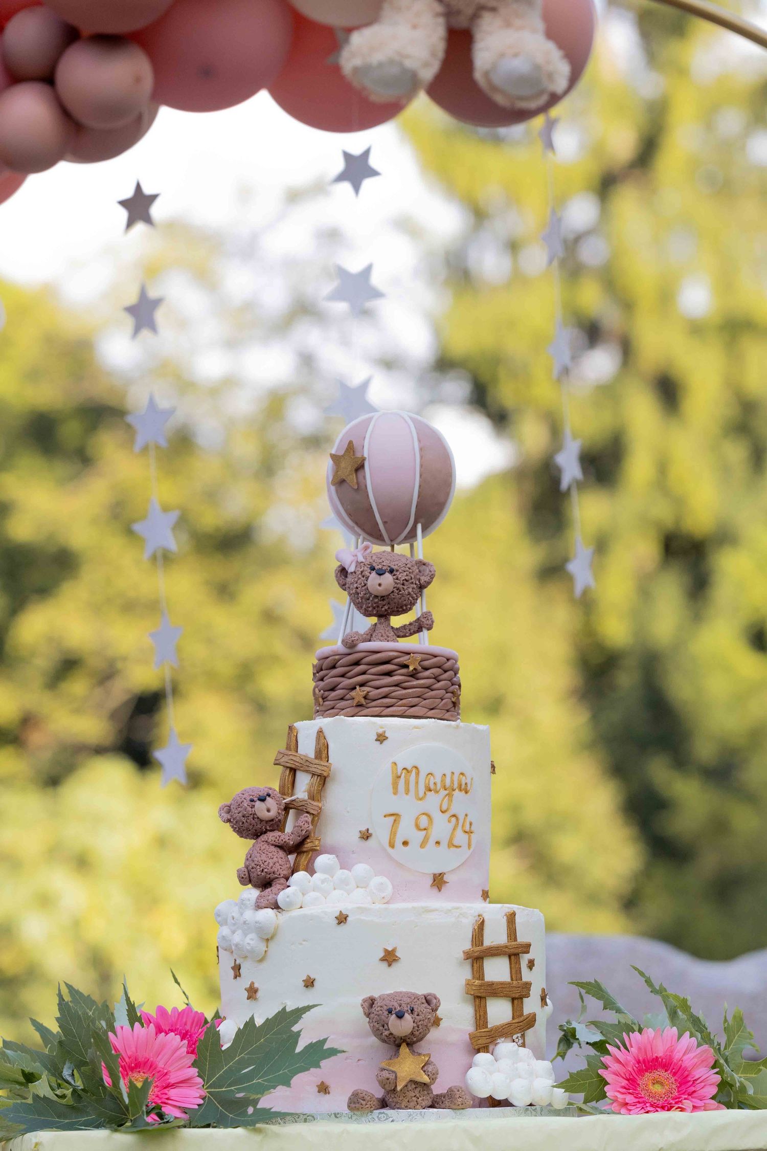 A white tiered cake decorated with teddy bear figures, stars, and pink flowers in an outdoor setting.