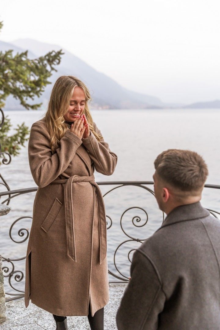 Man proposing to surprised woman in brown coat by scenic lake with mountains in background.