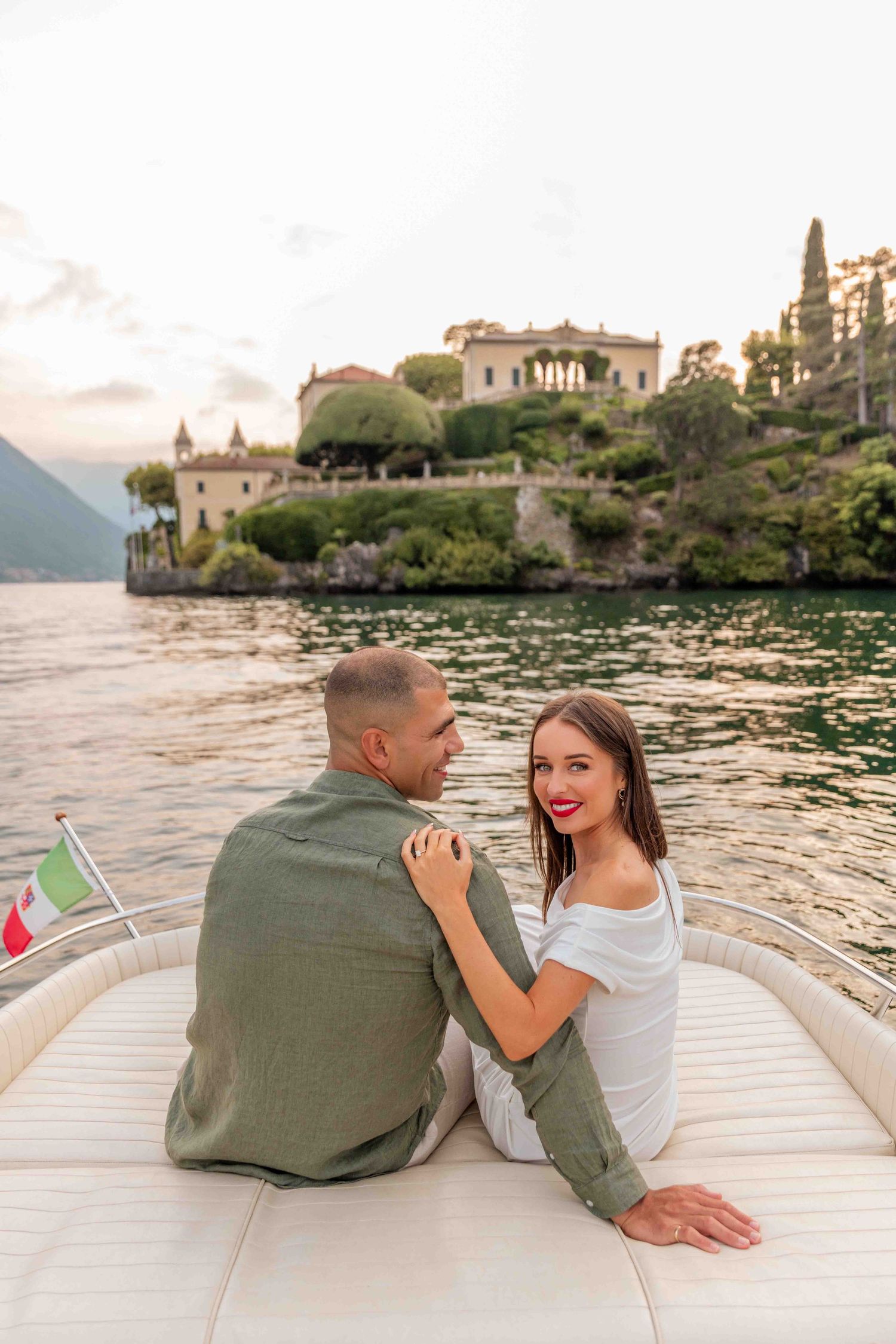 A couple relaxes on the cushioned deck of a boat with Italian villas visible along the shore at sunset.