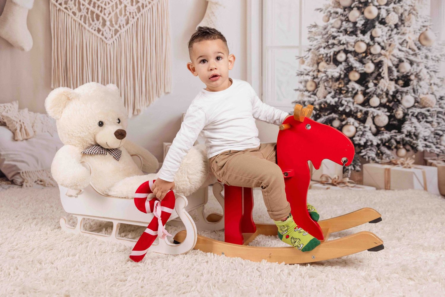 Child plays with stuffed toys near white Christmas tree and macrame wall hanging.