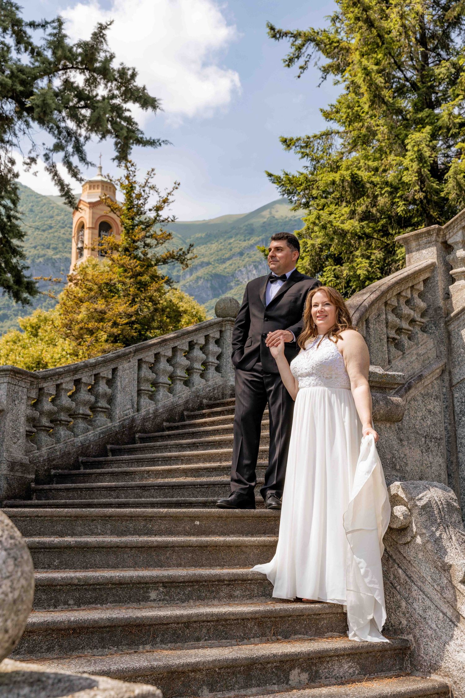A couple in wedding attire poses romantically on stone steps surrounded by lush greenery and mountains.