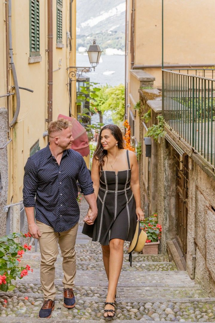 Couple walking through a charming cobblestone alley in Italy with red flowers and rustic architecture.