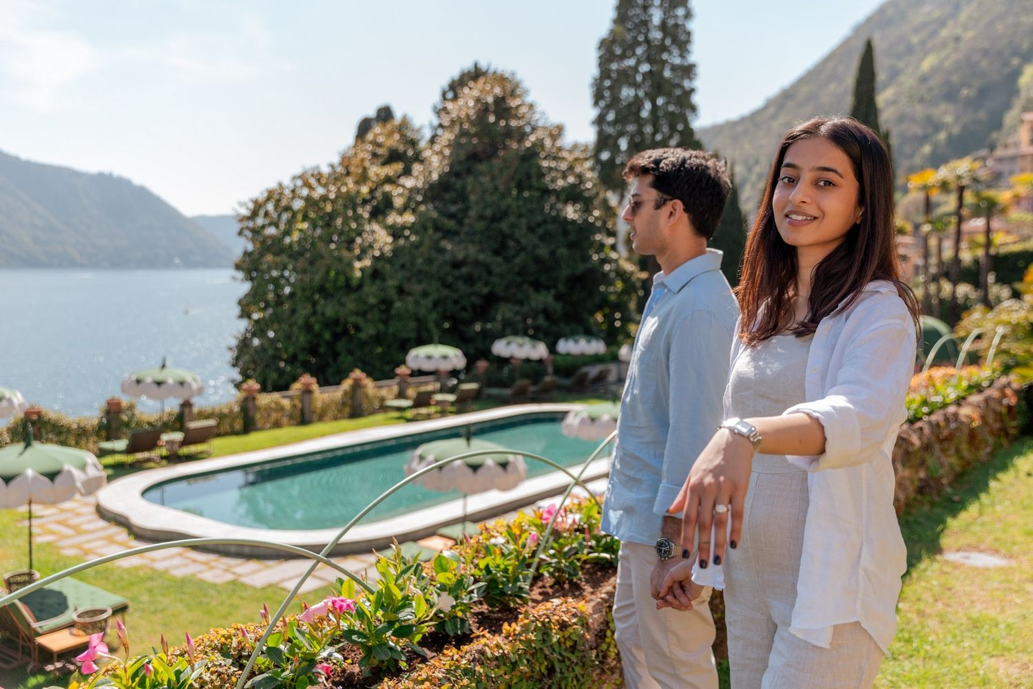 Couple holding hands by a luxury pool with scenic lake and mountain views on a sunny day.