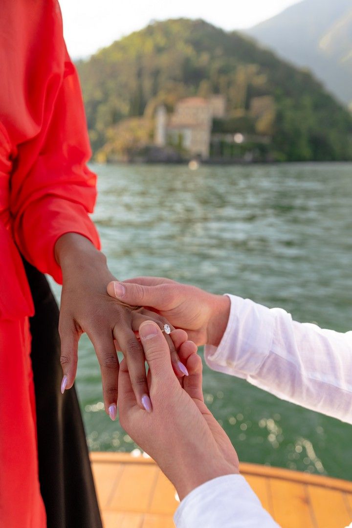 Person placing a diamond engagement ring on a woman's finger on a boat with a scenic lake and villa in the background.