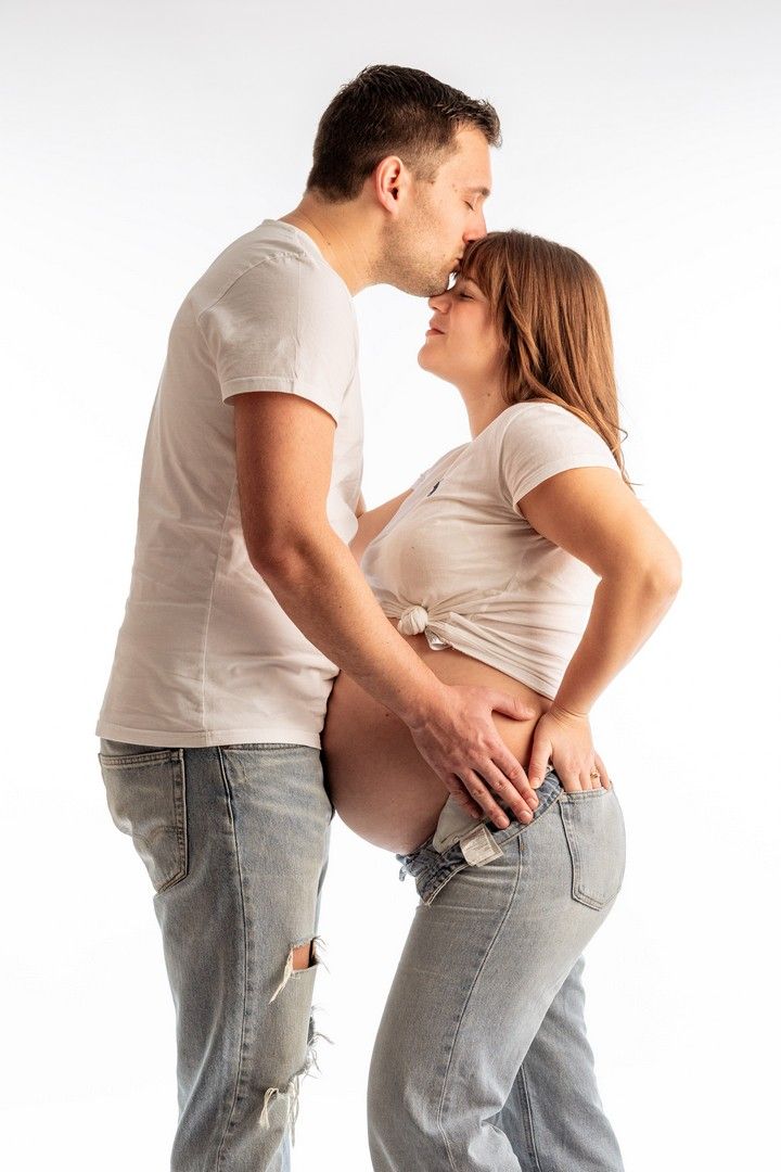 Pregnant couple sharing a kiss, both wearing white shirts and jeans, with man touching woman's baby bump.