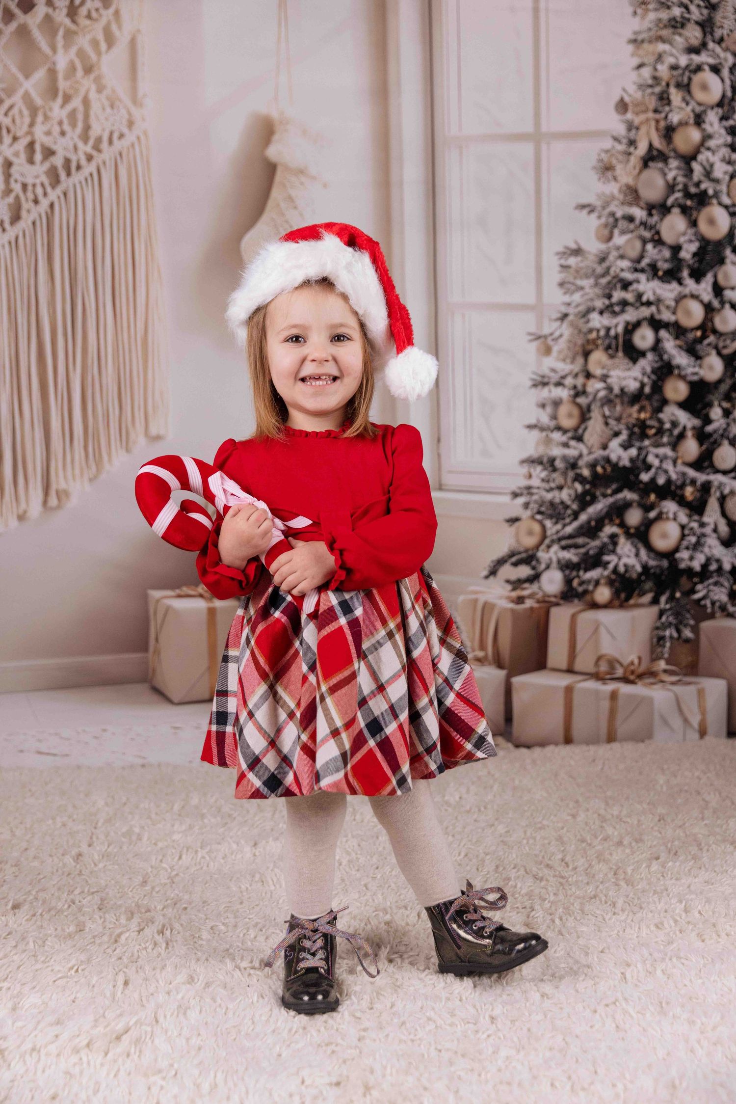 Child in red holiday outfit and plaid skirt sitting on white floor near Christmas tree.