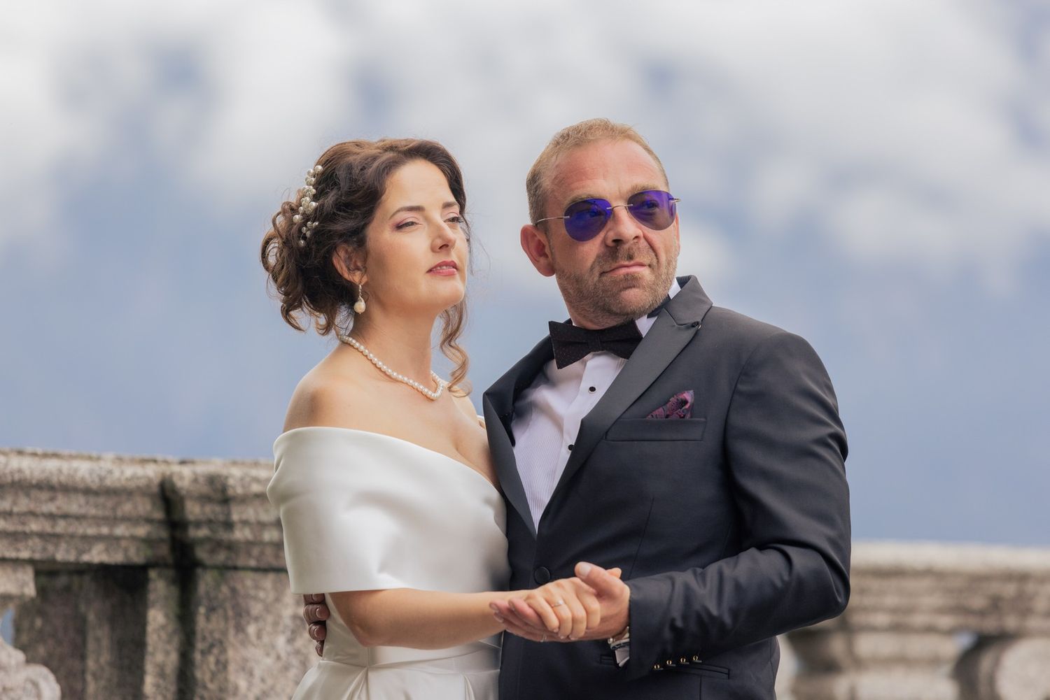 A couple poses together on a stone terrace, with the bride in white and the groom wearing sunglasses and a dark suit.