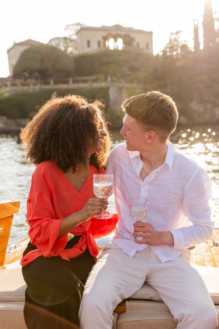Romantic couple sharing champagne on a boat at sunset, with a historic villa and sparkling water in the background.