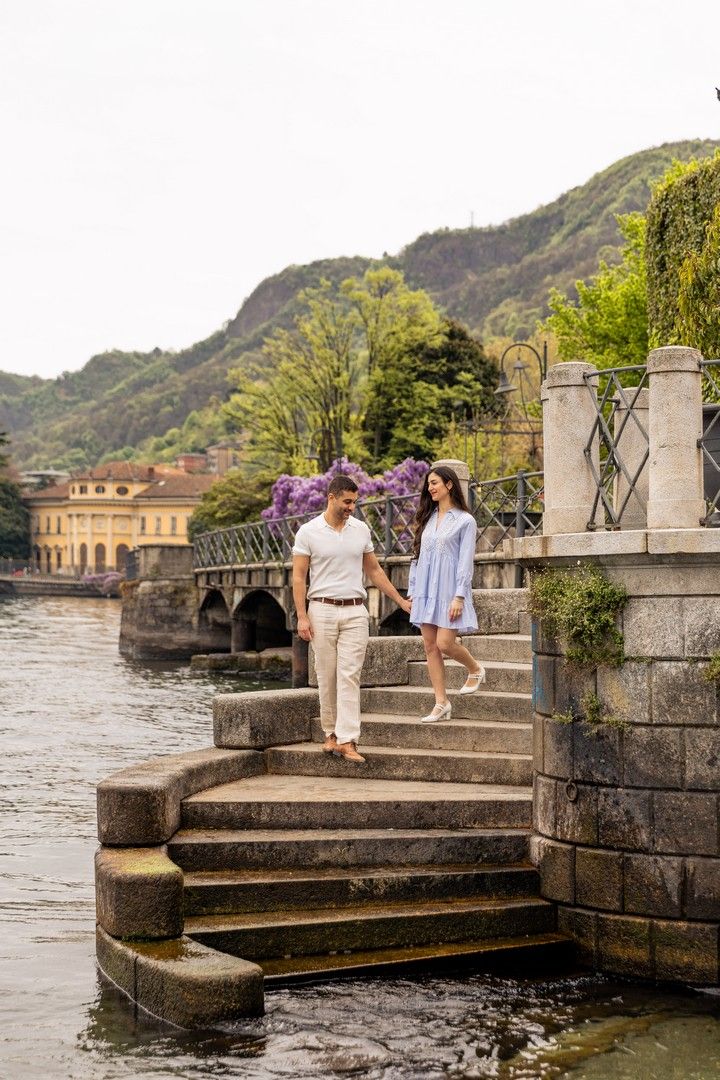 Couple holding hands walking down stone steps by a lakeside with lush green hills and blooming wisteria in background.