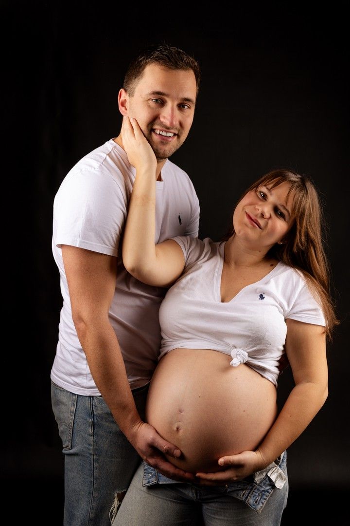 Happy couple expecting a baby, woman showing pregnant belly, both wearing white shirts, posing against black background.