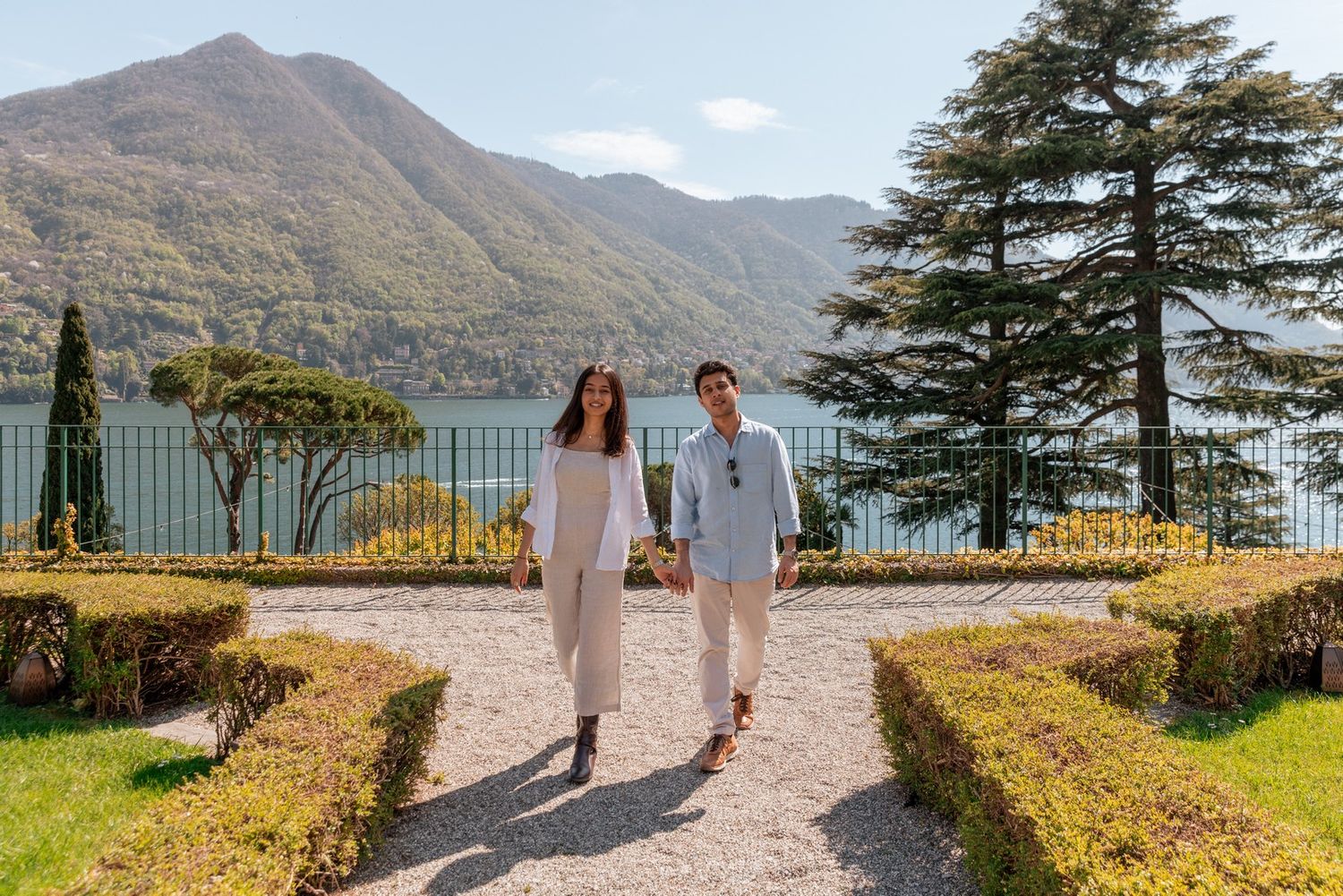 Couple walking hand in hand through formal garden with lake and mountain views at Villa del Balbianello, Lake Como.