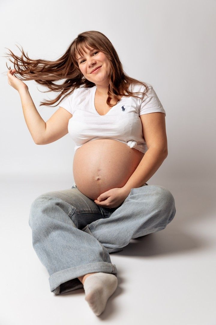 Smiling pregnant woman in white Polo shirt and jeans sitting cross-legged on floor with hair flowing.