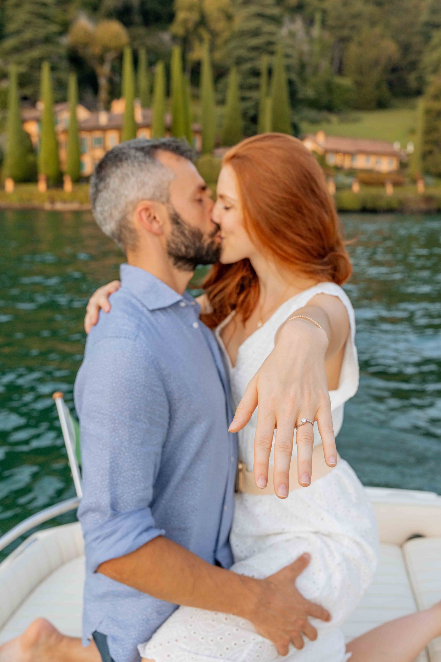 A romantic couple shares intimate kisses while sitting on a boat on a scenic lake with trees in the background.