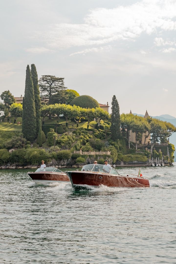 Wooden boats cruise across a lake with Italian cypress trees and villas dotting the scenic shoreline.