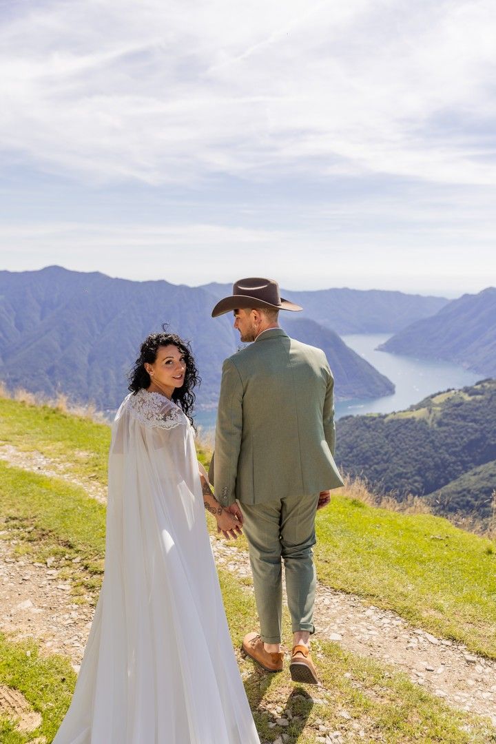 A couple walks hand in hand along a mountain trail overlooking a scenic lake and mountains in the distance.