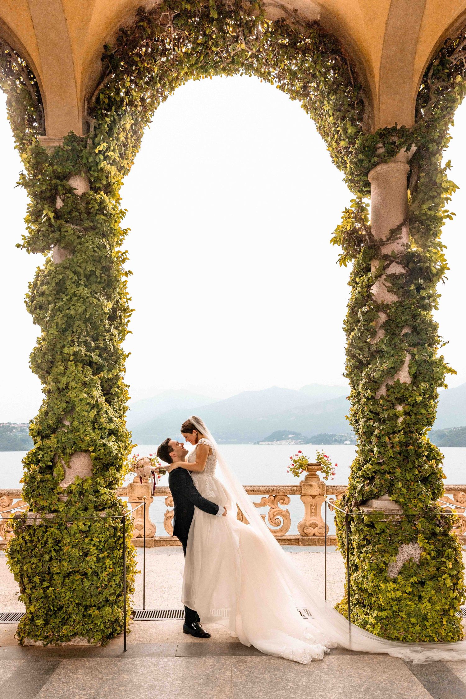 Romantic couple poses in a scenic archway covered with lush greenery overlooking Lake Como at sunset.