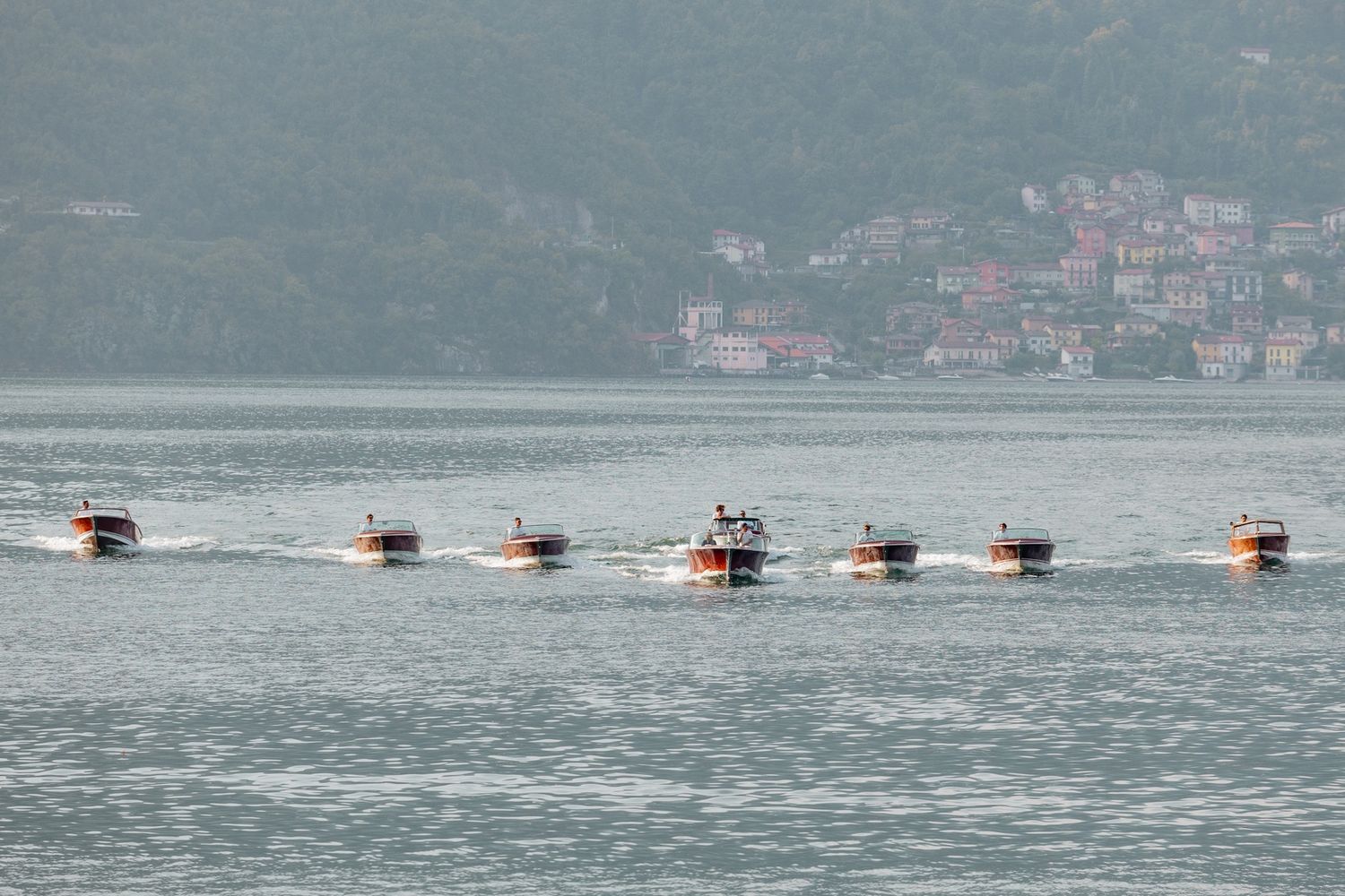 Seven classic Riva boats race in formation across a mountain-lined lake.