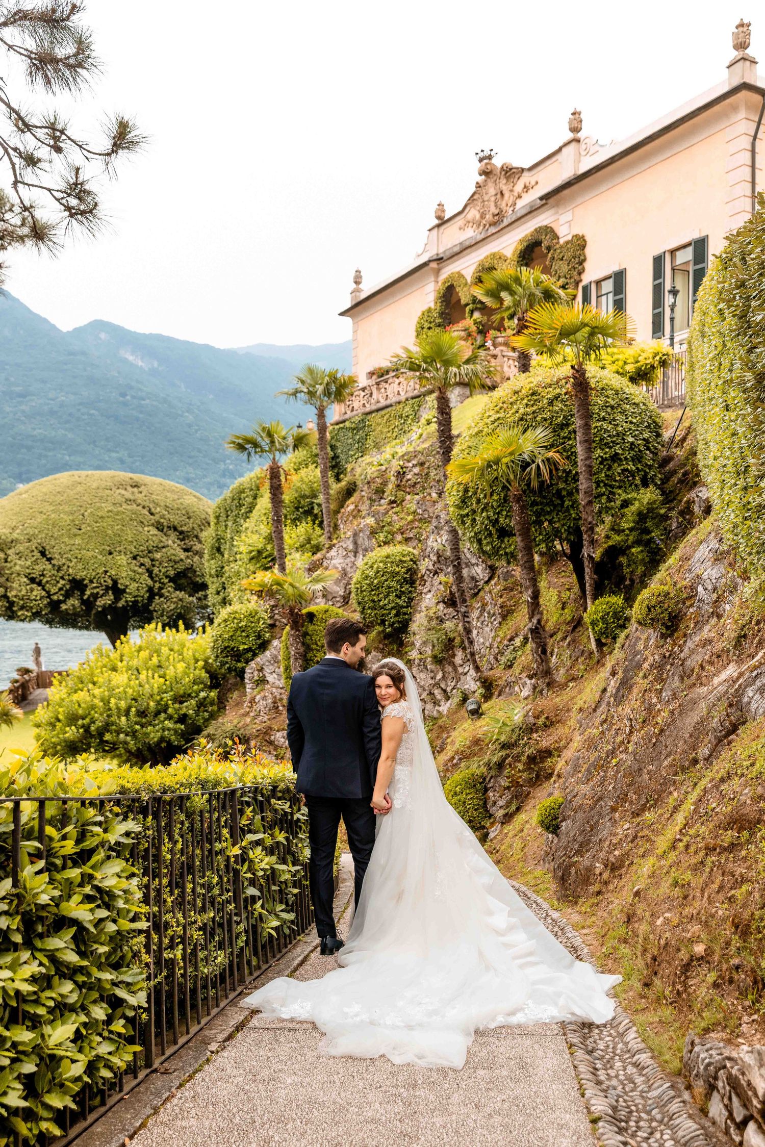 A couple stands on a scenic pathway at Villa del Balbianello overlooking Lake Como with mountains in the background.