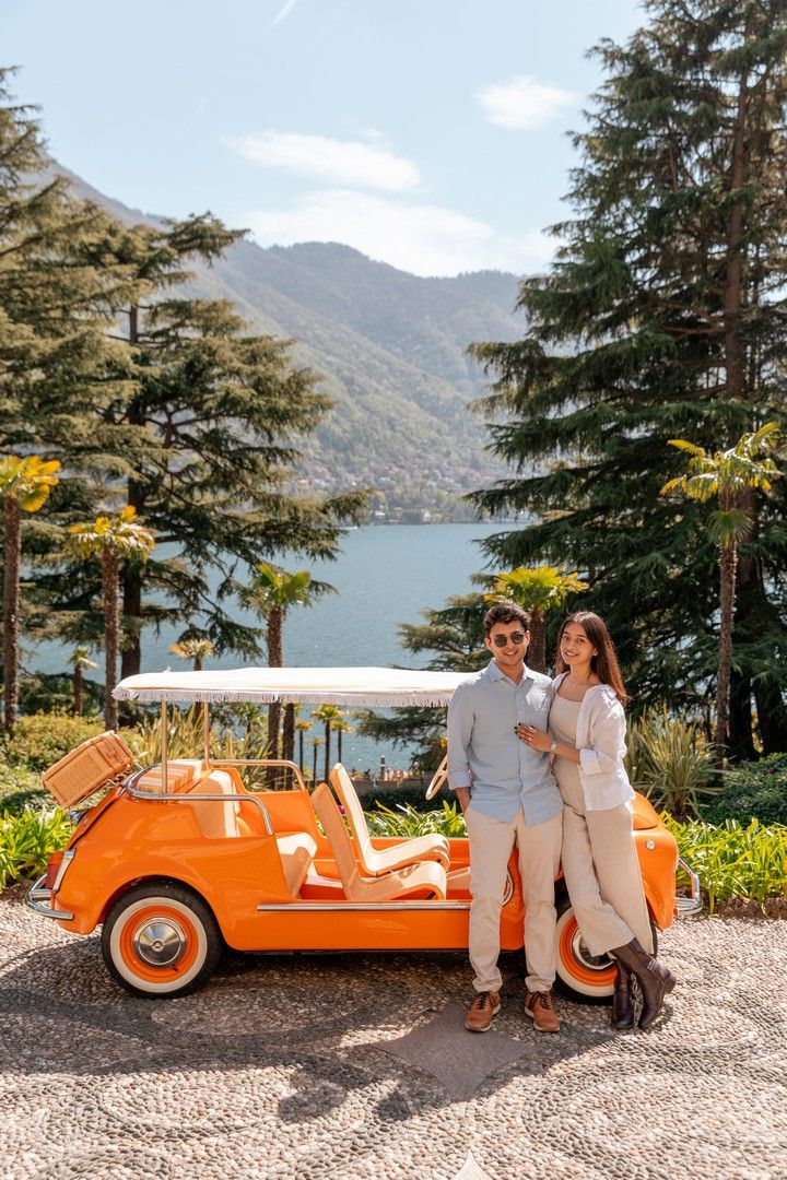 Couple posing by a vintage orange car at Lake Como, Italy, with lush trees and mountains in the background.