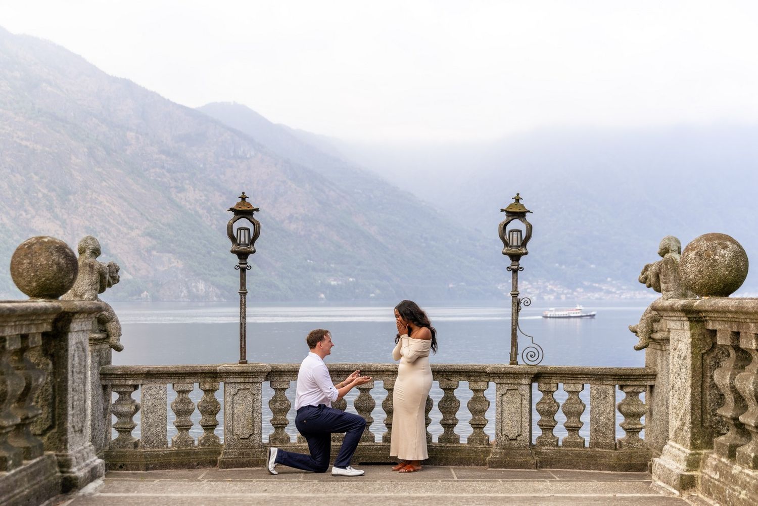 Man proposes to woman on ornate stone terrace overlooking Lake Como with mountain backdrop.