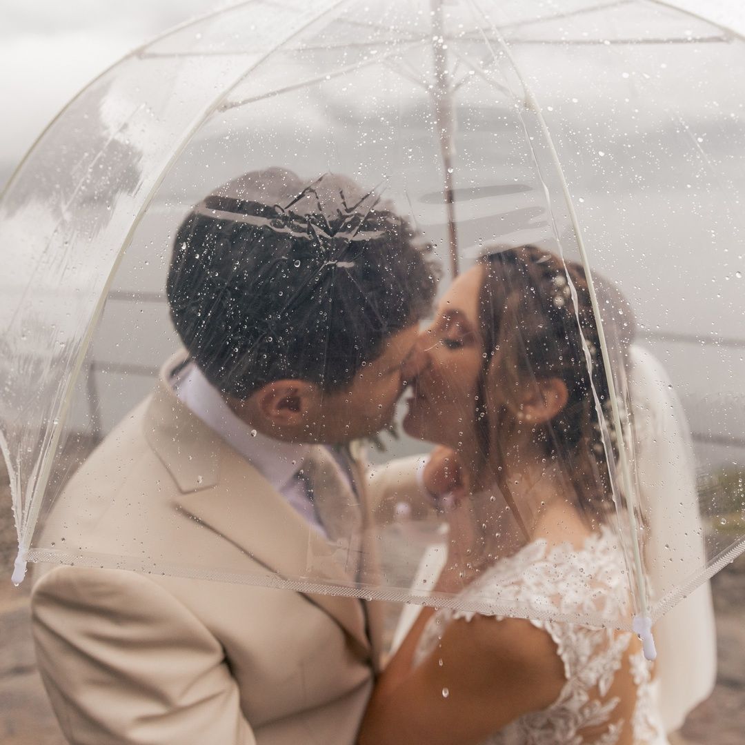 Romantic wedding photo of couple sharing intimate moment under clear umbrella in rain.