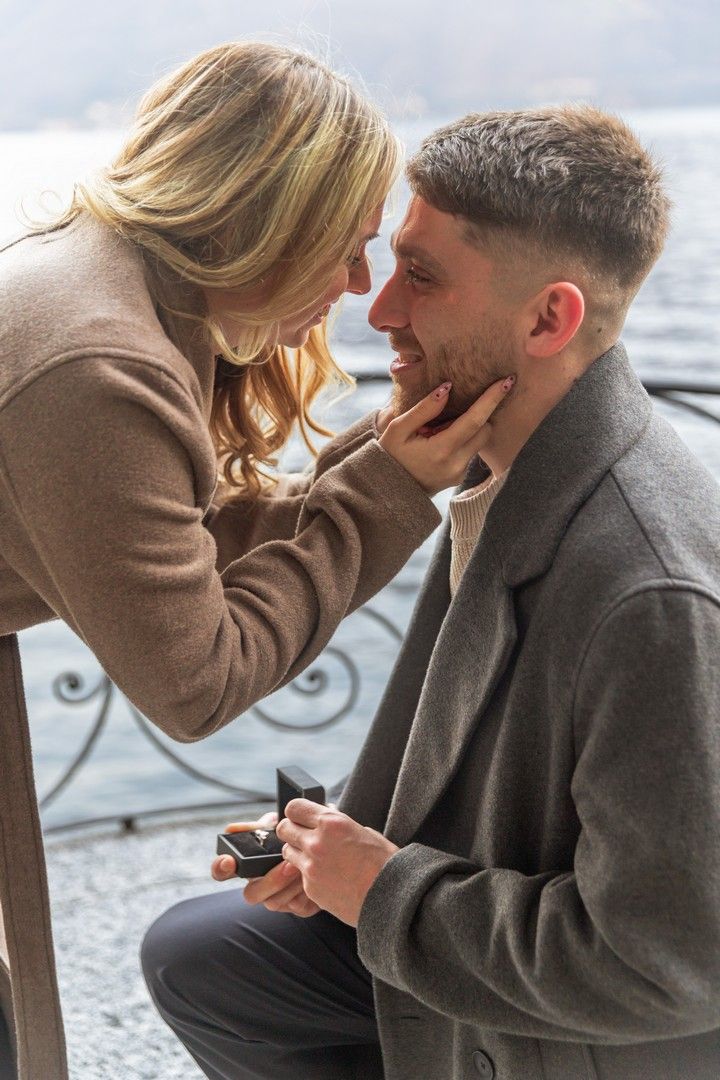 Man kneeling outdoors holding engagement ring box while woman tenderly cups his face near a scenic lakeside.