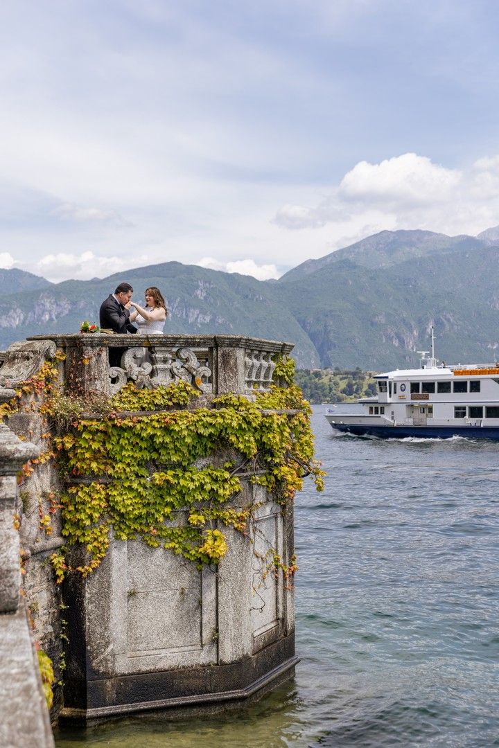 A romantic lakeside terrace with stone railings and yellow flowers overlooking mountains and a boat in the distance.