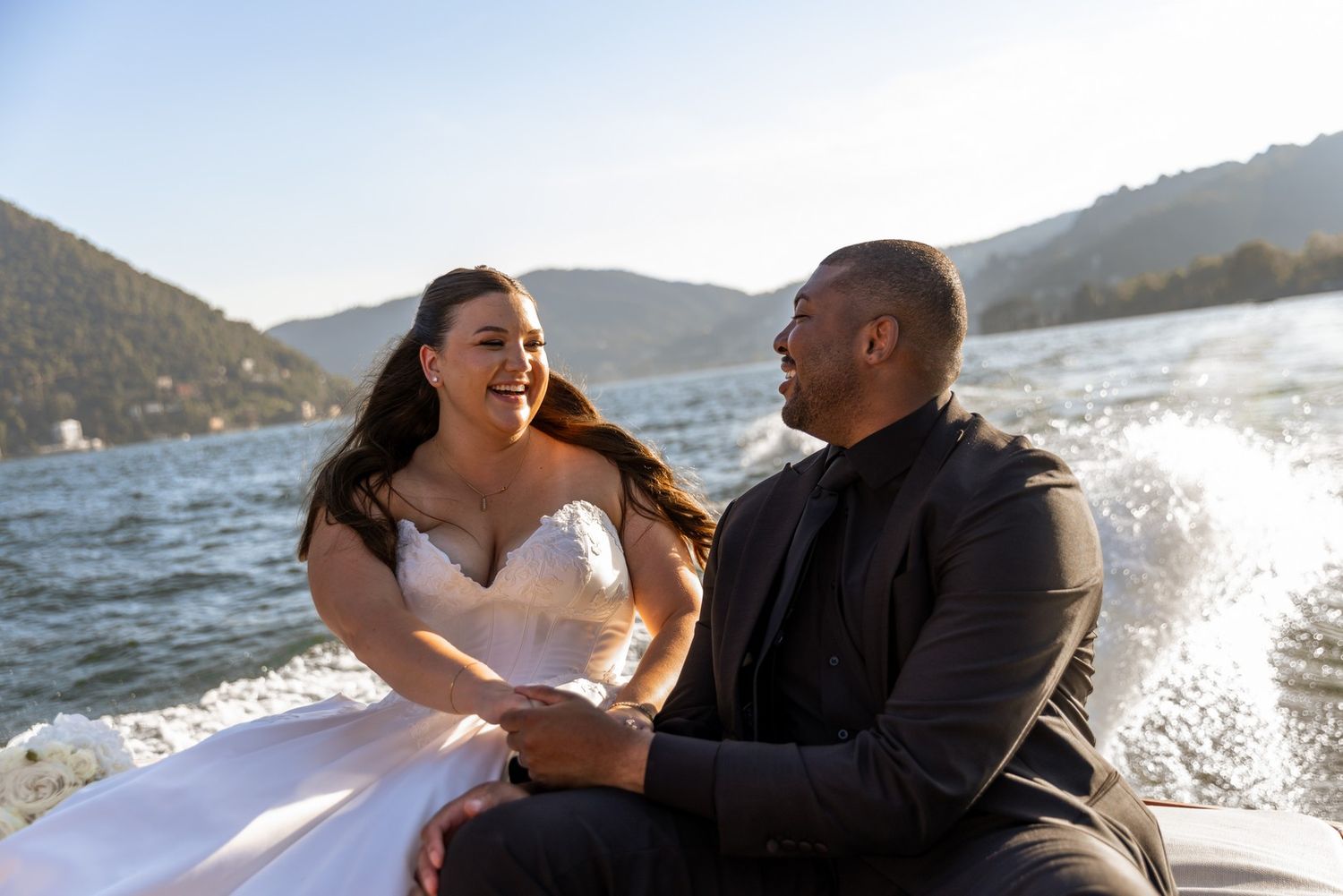A couple shares a romantic moment on a boat with sunlight sparkling on the water and mountains in the background.