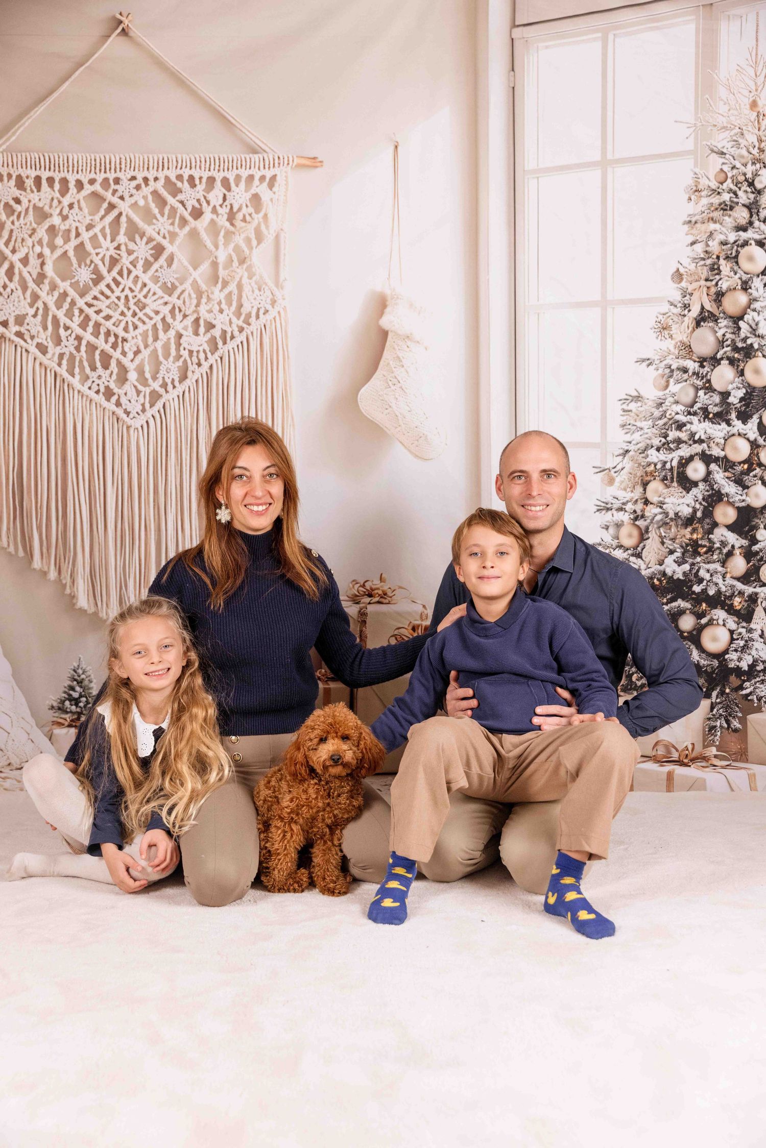 Family in matching navy outfits poses with brown dog against Christmas backdrop with macrame decor.