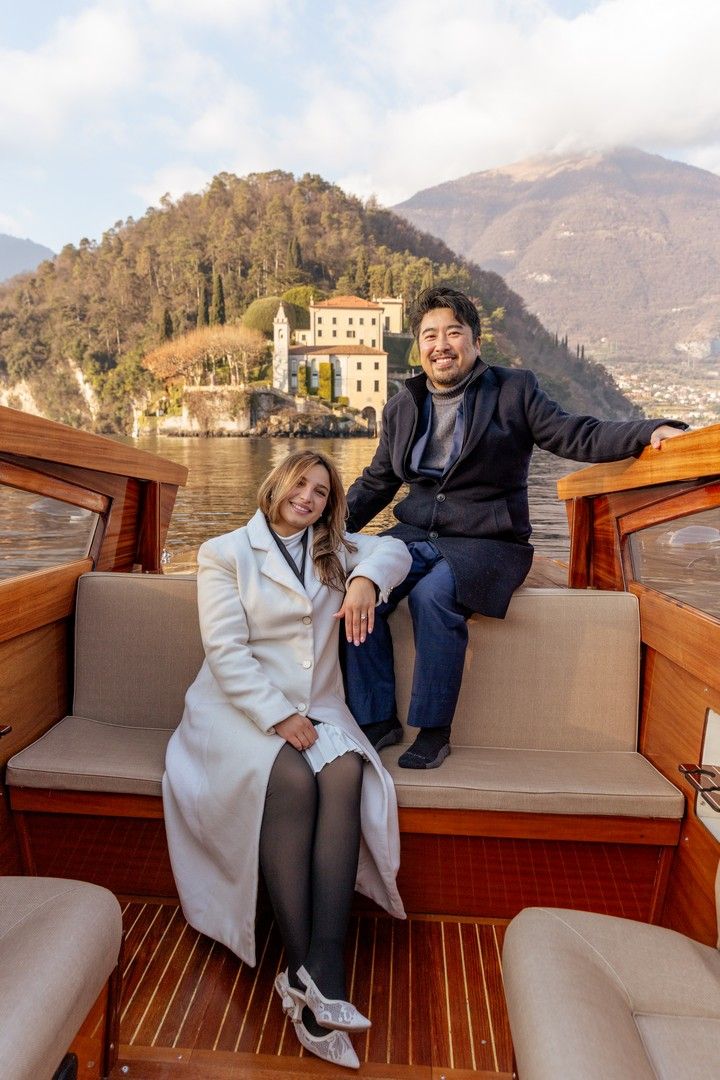 Smiling couple sitting on elegant wooden boat with Villa del Balbianello on Lake Como behind them.