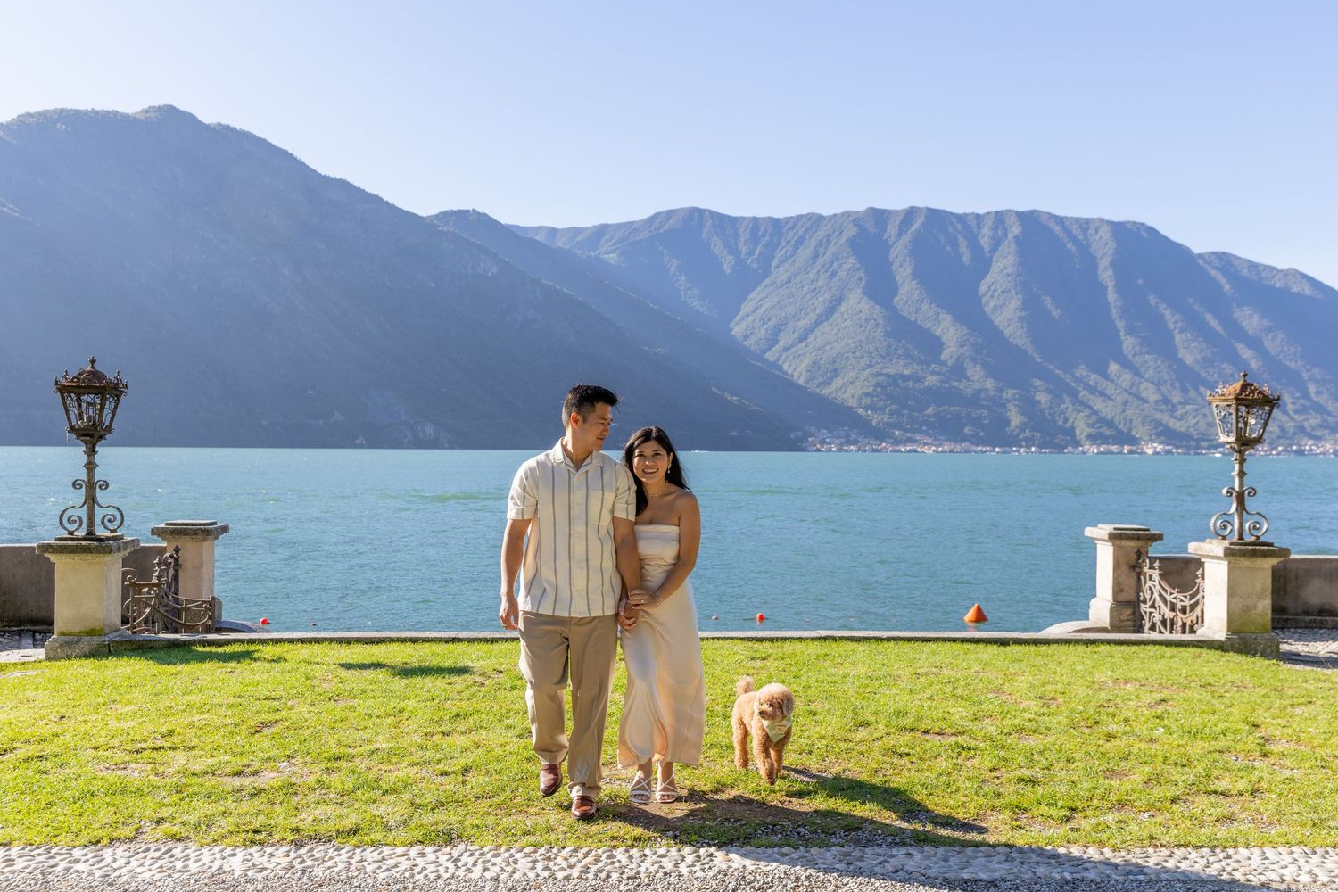 A couple enjoys a scenic mountain lake view from a grassy overlook on a sunny day.