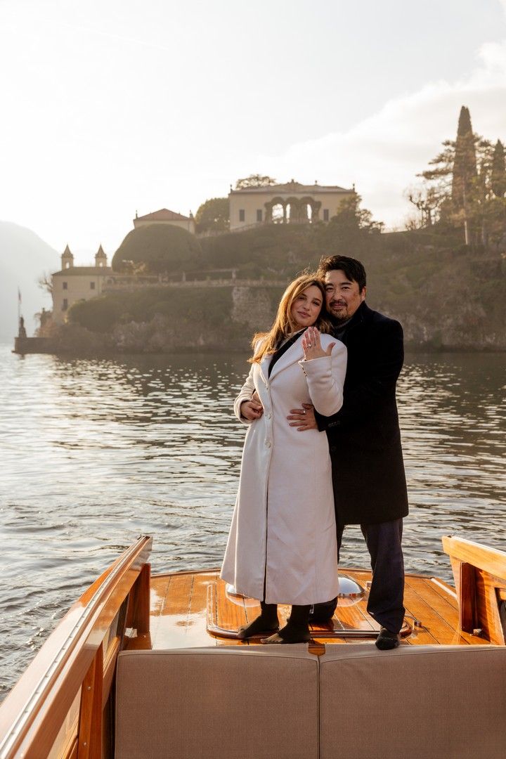 Happy engaged couple embracing on a wooden boat at Lake Como with historic villa backdrop.
