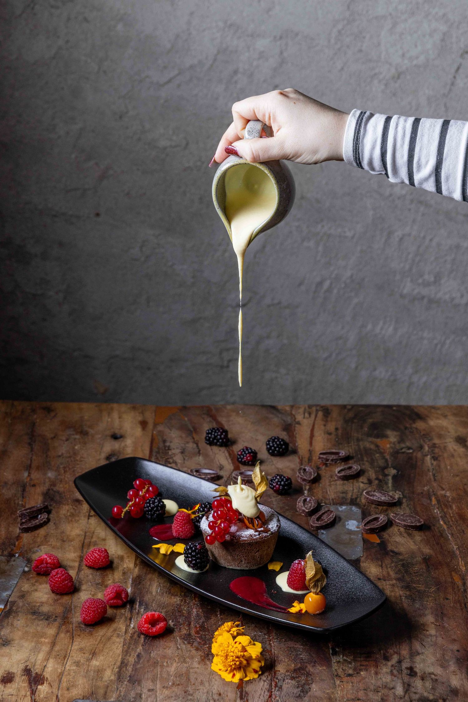 Drizzling sauce over a dessert plate decorated with fresh berries and edible flowers.
