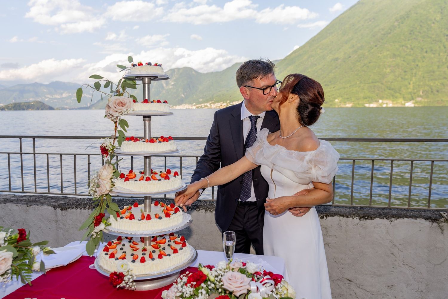 A wedding couple shares a tender moment beside their elegant four-tier cake decorated with fresh berries by a lakeside terrace.