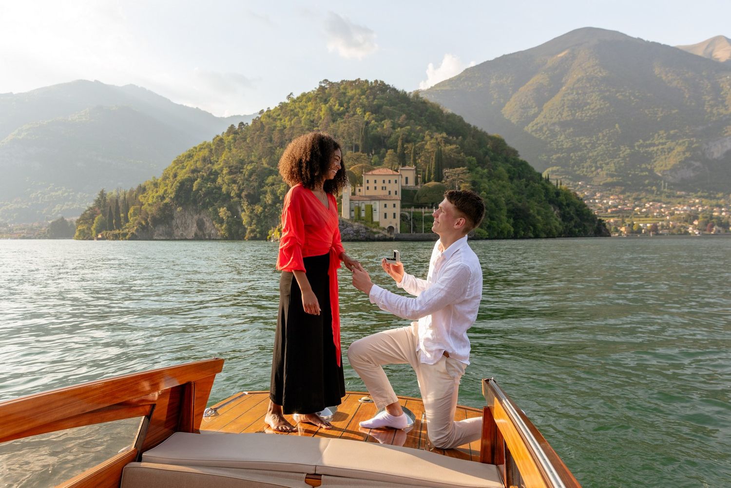 Man proposes to woman on a wooden boat on Lake Como, Italy, with mountain and villa backdrop.
