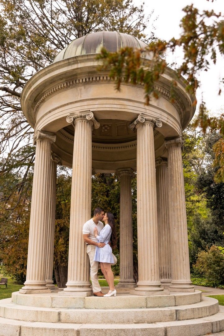 Couple embracing romantically under a classical stone rotunda with Ionic columns in a lush park setting.