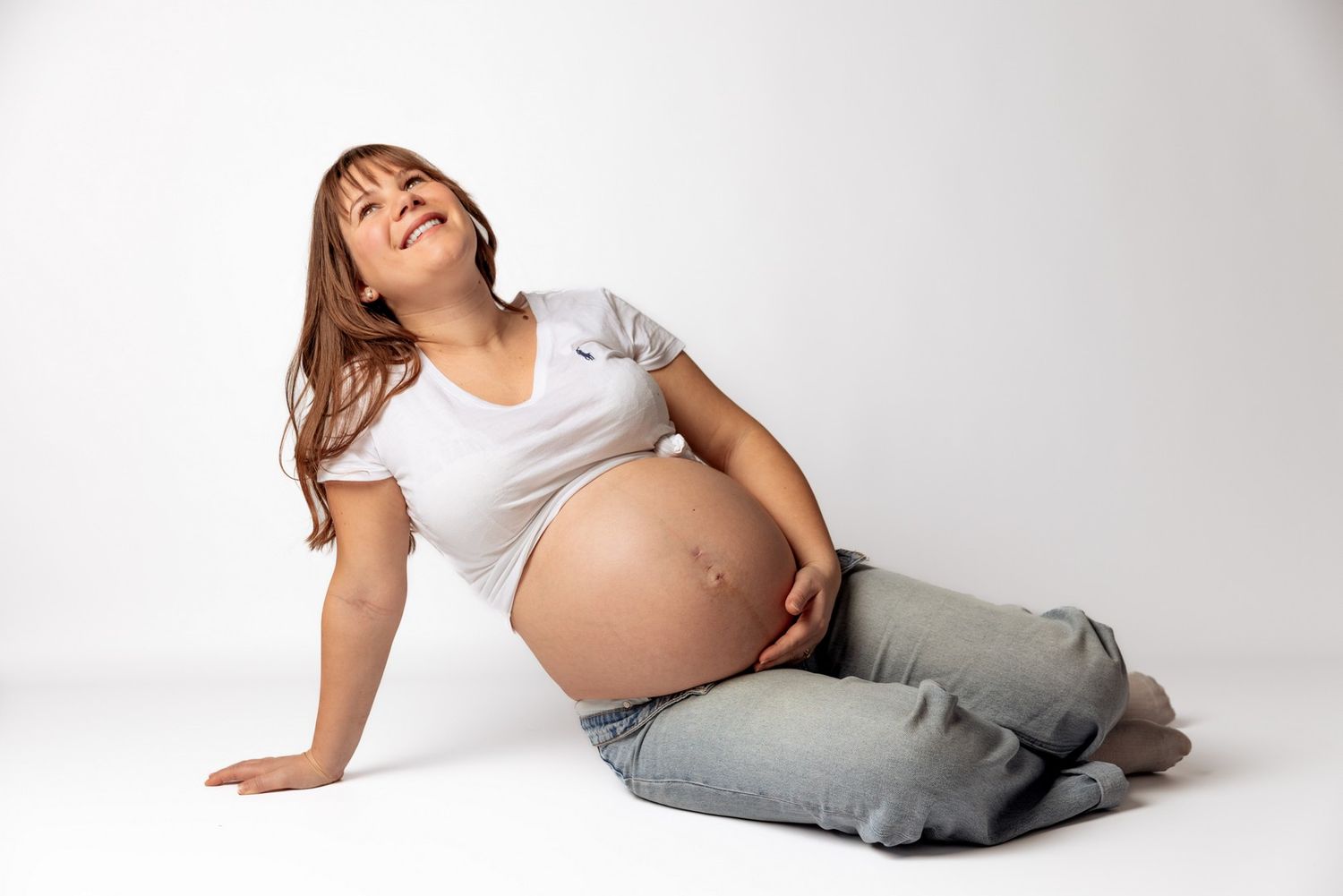 Smiling pregnant woman in white t-shirt and gray pants sitting on floor cradling her baby bump.